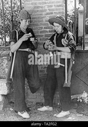 Foto von zwei Republikanische Kämpferinnen der Arbeitermiliz Mittagessen an der Vorderseite der Sierra Guadarrama nördlich von Madrid in kurz nach dem Ausbruch des Spanischen Bürgerkrieges im August 1936. Die beiden Frauen, die Bänder sind mit Karabiner bewaffnet. Ihre Mahlzeit besteht aus Konserven. Sie tragen alpargatas, leichte Baumwolle Schuhe mit geflochtenem Hanf Sohlen. Die zwei schlanken gegen eine Wand mit einem Vorgarten. Stockfoto