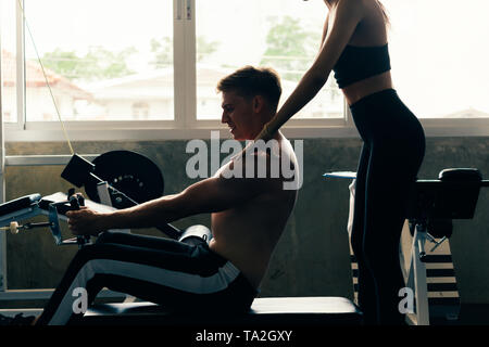 Jungen kaukasischen Mann im weißen Hemd an einer Turnhalle, harten Trainings ziehen Gewichte im eingesetztes Reihe Maschine mit Unterstützung von weiblichen Personal Trainer Stockfoto