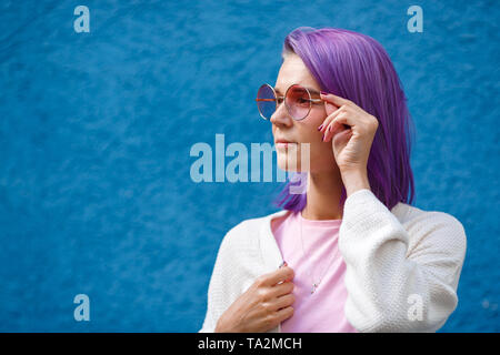 Schöne junge Mädchen mit lila Haaren in rosa Brille auf dem blauen Hintergrund schaut auf die Seite, weisse Jacke, Hand hält Gläser, Rot, Maniküre Stockfoto