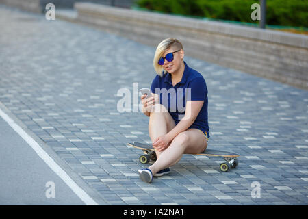 Stilvolle Mädchen von großen Größen, modischer Haarschnitt im Mirror Gläsern, sitzen auf einem longboard vor dem Hintergrund der Gebäude, die das Telefon Stockfoto