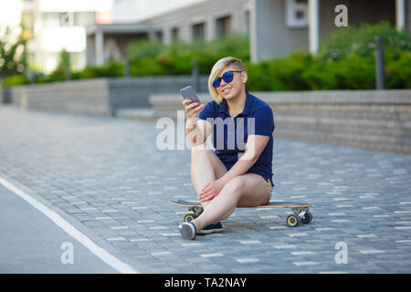 Stilvolle Mädchen von großen Größen, modischer Haarschnitt im Mirror Gläsern, sitzen auf einem longboard vor dem Hintergrund der Gebäude, die das Telefon Stockfoto