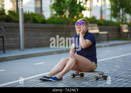 Stilvolle Mädchen von großen Größen, modischer Haarschnitt im Mirror Gläsern, sitzen auf einem longboard, Telefonieren, lächelnd Stockfoto