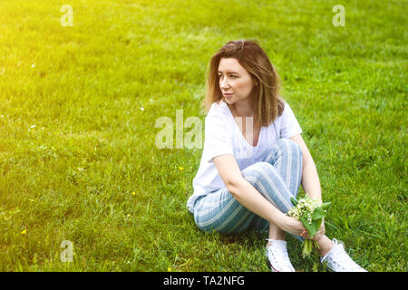 Stilvolle junge Frau mit Blumen und sitzen auf grünem Gras im sonnigen warmen Tag Stockfoto