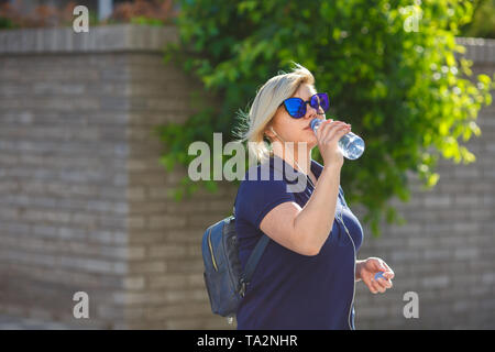 Stilvolle Mädchen plus Größe in blauen T-Shirt und Sonnenbrille Getränke Wasser aus einer Flasche, hört zu, Kopfhörer, vor dem Hintergrund einer Wand und Holz Stockfoto