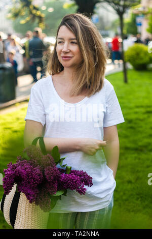 Stilvolle Frau mit Stroh Beutel mit einem lebendigen Haufen lila Blüten. Helle grüne Gras Hintergrund. Stockfoto