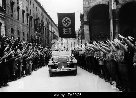 Adolf Hitler Laufwerke in einem offenen Wagen entlang der Residenzstraße in München. Rechts und links der Straße ist der Feldherrenhalle und der Residenz. Zwischen den beiden Gebäuden ist eine Flagge mit Hakenkreuz und ein Dreibettzimmer siegrune. Undatiertes Foto. Stockfoto