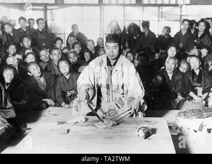 Ein Koch im traditionellen Kimono sitzt vor einem Schneidbrett in Tokio und schneidet einen Fisch mit einem langen Messer und essstäbchen vor einem Publikum. Stockfoto