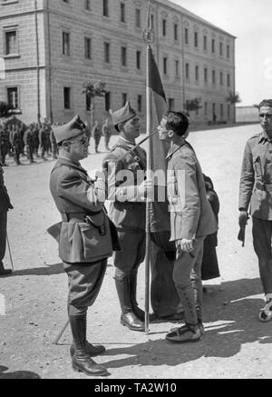 Undatiertes Foto von einer Vereidigung von Spanischen Nationalen Rekruten in Salamanca nach Ausbruch des Spanischen Bürgerkriegs im Jahre 1936. Das Bild zeigt eine Teniente (Leutnant), auf der linken Seite mit Sonnenbrillen das Tragen einer Uniform, der ein Schwert über einen zu rekrutieren (rechts, mit einheitlichen Rock und alpargatas, einfachen Espadrilles), um ihn zu küssen. Zwischen ihnen steht eine Alferez (Leutnant) mit der regimental Standard. Die Soldaten warten auf ihre Vereidigung beim Stehen in der Linie. Im Hintergrund, Soldaten und die kaserne Gebäude. Stockfoto