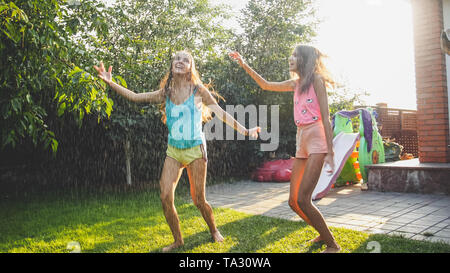 Foto von zwei glücklich lachend Schwestern in nassen Kleidern tanzen Wassertropfen aus Gartenschlauch im Garten. Familie Spiel und Spaß im Freien an Stockfoto