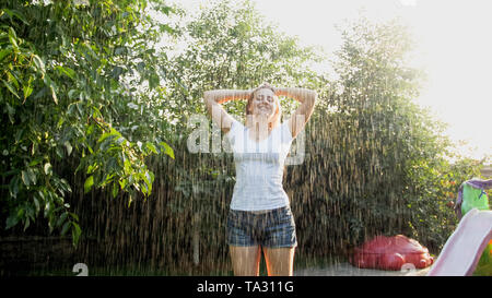 Porträt der Schönen lächelnde Frau in nassen Kleidern genießen Sie warmen Regen im Haus Garten im Hinterhof bei Sonnenuntergang. Mädchen spielen und Spaß im Freien an Stockfoto