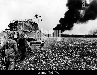 Ein deutscher Kampfpanzer VI (Tiger) in einer Wiese in Litauen. Hinter, mehrere Infanteristen unter dessen Abdeckung. Im Hintergrund, der Rauch von einem brennenden Haus. Foto der Propaganda Firma (PK): Kriegsberichterstatter Slickers. Stockfoto