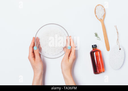 Die Ansicht von Oben weibliche Hände halten Schüssel mit Meersalz neben Flasche Rosmarin aromatische Öl- und Bimsstein auf weißem Hintergrund. Flach Zusammensetzung der Stockfoto