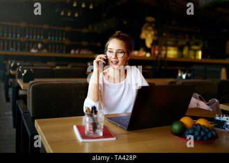 Junge Freiberufler arbeiten mit Laptop und Gespräch am Handy mit Client im Cafe. Hübsche asiatische kaukasischen gemischten Rennen Business woman in Gläsern Verhalten Stockfoto