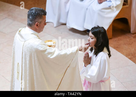 Zagreb, Kroatien - 5. Mai 2019: Der Priester gibt Erstkommunion für die Kinder in weißen Kleidern während der Kommunion in der Kirche von Alle gekleidet Stockfoto