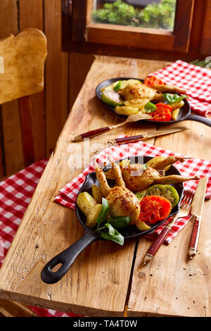 Tabelle für zwei mit Portionen von gebratene Wachtel und frischem Gemüse auf Gusseisen skillets in einem rustikalen Taverne oder im Restaurant Stockfoto