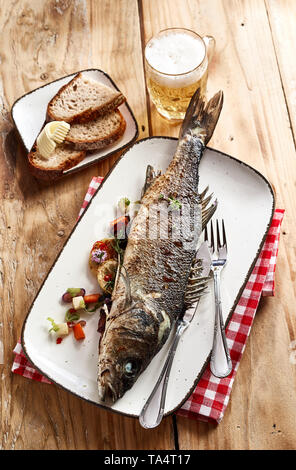 Ganze Ofen gegrillten Wolfsbarsch mit einem kalten Bier mit Roggen Brot auf einem rustikalen oder Vintage Holz Tisch serviert gesehen hohen Winkel Stockfoto