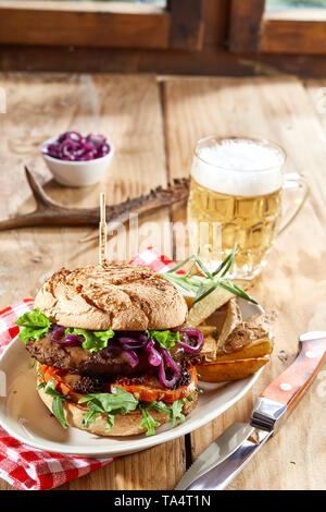 Wild Wild Beef Burger mit Salat Beilagen auf einem Teller mit Kartoffelecken und eine kalte schaumiges Bier auf einer rustikalen Tisch serviert, Seitenansicht für Menü Stockfoto