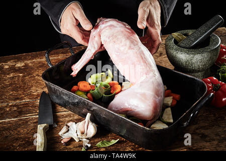 Der Küchenchef bereitet eine enthäutete gereinigt Wilde Kaninchen Schlachtkörper für Braten in einer Pfanne mit Lauch, Karotten und Tomaten und verschiedene Gewürze Stockfoto