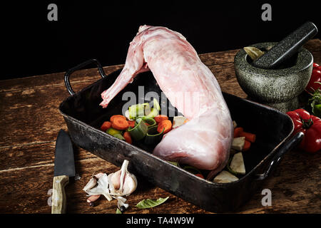 Raw gereinigt Wilde Kaninchen warten zum Braten in einer Pfanne mit gehackter Lauch, Karotten, Knoblauch, Kräuter und Tomaten auf eine rustikale Küche Tabelle gehäutet Stockfoto