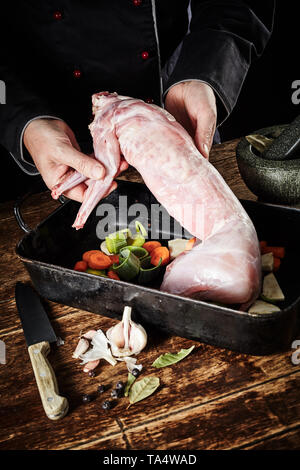Der Küchenchef bereitet Wilde Kaninchen zum Rösten in einer rustikalen Küche Anzeige der gereinigt enthäuteten Schlachtkörpern zur Ansicht in einem Bräter mit gewürfelten Gemüse Stockfoto