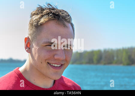 Halbe Umdrehung büste Portrait von einem jungen Mann im roten T-Shirt, stehen draussen auf dem See oder Fluss. Auf Kamera und lächelnd Stockfoto