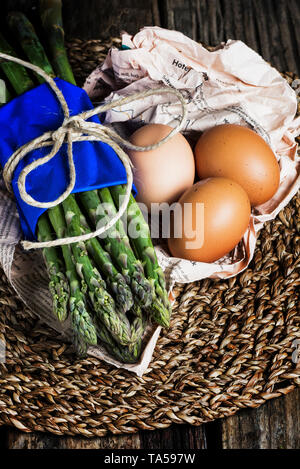 Bündel Spargel mit Eiern auf den Mittelpunkt, mit blauem Papier gebunden, dunkle Stimmung Holz- Hintergrund Stockfoto
