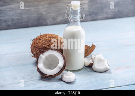 Kokosmilch in eine Flasche in der Zusammensetzung mit einem offenen Coconut mit weißem Fleisch auf einem blauen Hintergrund aus Holz. Organische gesunde Ernährung vegan Produkt verwenden Stockfoto