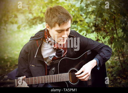 Ein junger Mann in einem schwarzen windbreaker und eine karierte Schal spielt eine schwarze Gitarre, im Park sitzen an einem sonnigen Sommertag. Stockfoto