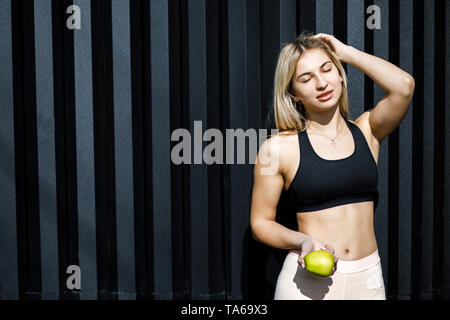 Eine junge, sympathische blonde Frau aktiv Ausgabe Zeit im Freien, einen grünen Apfel. Das Mädchen ist in Sportkleidung gekleidet. Zeit für eine gesunde sna Stockfoto