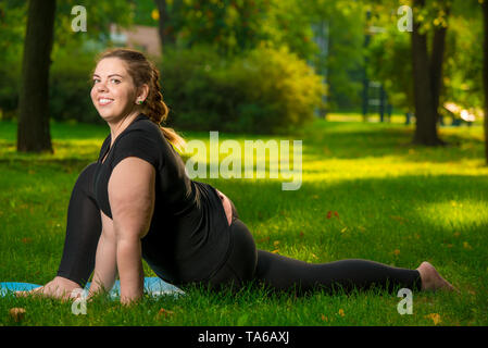 Junge flexible und schöne Frau plus Größe im Park auf der Wiese führt Dehnübungen Stockfoto