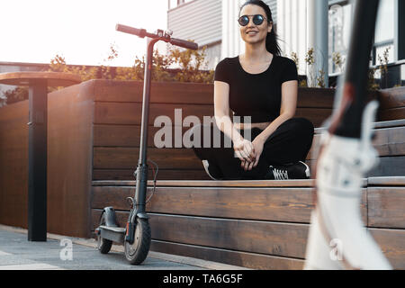 Junge schöne Frau sitzt auf der Holzbank am schönen Stadtbild Hintergrund in der Nähe von elektrischen Kick scooter. Stockfoto