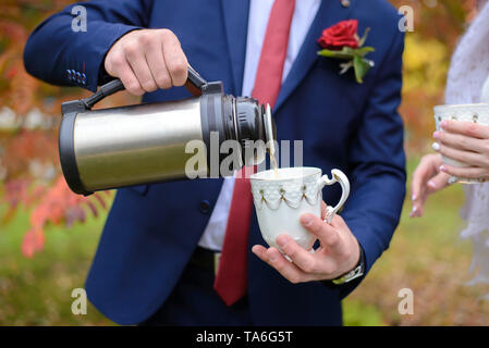 Der Bräutigam gießt Tee aus einer Thermoskanne in die weiße Tasse zu seiner Braut Stockfoto