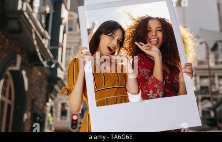 Zwei Frauen tragen bunte Kleidung stehen im Freien mit einem Bilderrahmen und eine lustige Gesichtsausdrücke. Schöne weibliche Freunde genießen, Stockfoto