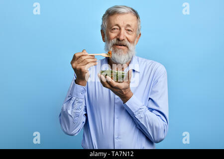 Happy bärtiger Mann im blauen Hemd Essen Salat, health care Konzept. plus alt Mann hält eine Schüssel mit Salat und Gabel, gesundes Essen, Mann führt Gesundheit Stockfoto