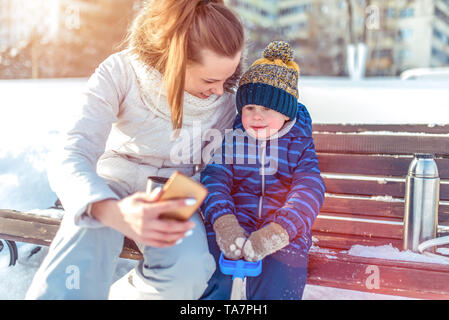 Eine Frau Mutter mit Kind ist auf einer Bank im Winter in der Stadt fotografiert, in warme Kleidung, ein kleiner Junge von 3 Jahren in einem blauen Overalls und ein Stockfoto