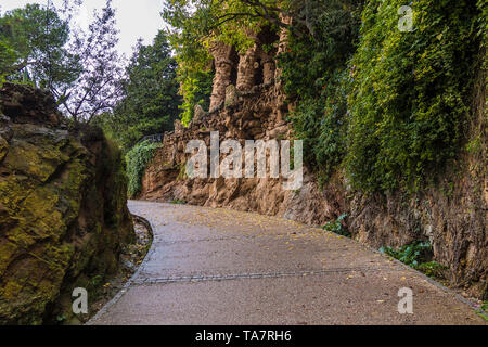 Ein Fußweg zwischen zwei Steinmauern mit üppiger Vegetation im Park Güell in bewölkten Herbst Tag, Barcelona, Katalonien, Spanien Stockfoto