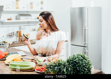 Ziemlich stylish girl Holding schneiden Avocado in der Nähe von Tisch in der Küche Stockfoto