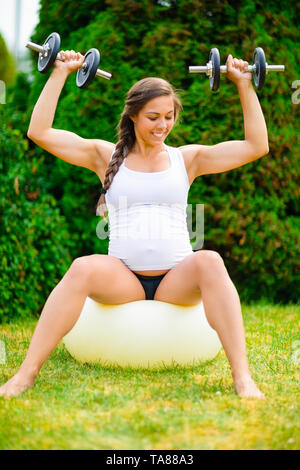 Zuversichtlich schwangere Frau Hanteln auf Yoga Ball im Park Stockfoto