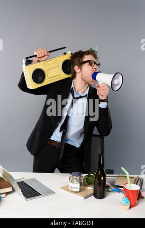 Wütend Geschäftsmann am Arbeitsplatz in der Partei cap Holding vintage tape recorder und schreiend in Megafon auf grauem Hintergrund Stockfoto