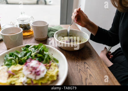 Frau das Essen von frischem Gemüse detox Suppe aus Brokkoli mit mit Kürbiskernen. Im Vordergrund Toast mit Avocado und pochiertem Ei, Spinat, Salat. Stockfoto