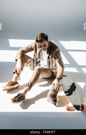 Stattlicher Mann mit adorable Waschbär sitzend im Sonnenlicht in der Nähe longboard Stockfoto