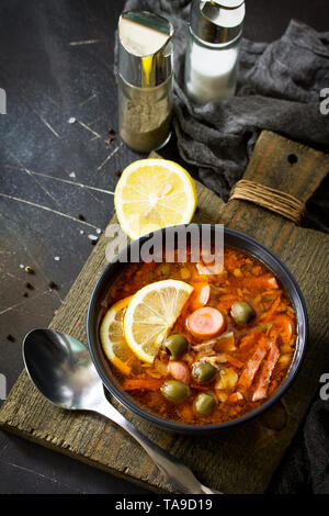 Traditionelle russische Suppe Solyanka mit Fleisch, Wurst, Gemüse, Kapern, Essiggurken und Oliven mit Zitrone auf schwarzem Hintergrund. Im rustikalen Stil. Freier Speicherplatz Stockfoto