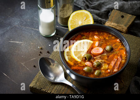 Traditionelle russische Suppe Solyanka mit Fleisch, Wurst, Gemüse, Kapern, Essiggurken und Oliven mit Zitrone auf schwarzem Hintergrund. Im rustikalen Stil. Freier Speicherplatz Stockfoto