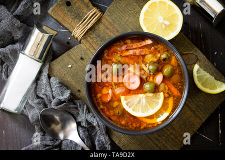 Traditionelle russische Suppe Solyanka mit Fleisch, Wurst, Gemüse, Kapern, Essiggurken und Oliven mit Zitrone auf schwarzem Hintergrund. Im rustikalen Stil. Ansicht von oben f Stockfoto