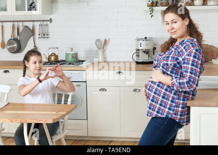 Zehn Jahre alte Tochter zeigen Herz für die werdende Mutter und großen Bauch, häusliche Küche Innenraum Stockfoto