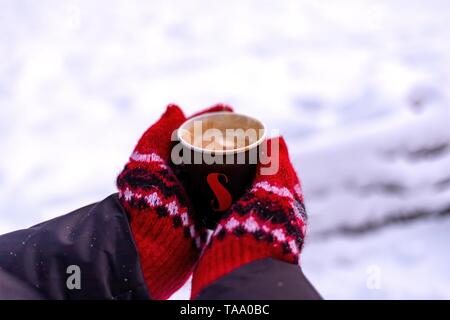 Frauenhände in einem roten Handschuhe halten eine Tasse Kaffee. Rückseite Blick auf die weiß verschneiten Hintergrund. Stockfoto