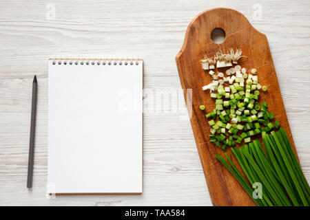 Gehackte Frühlingszwiebeln auf einem rustikalen Holzbrett, leere Notepad mit Bleistift über weiß Holz- Oberfläche, Ansicht von oben. Overhead, flach, von oben. Stockfoto