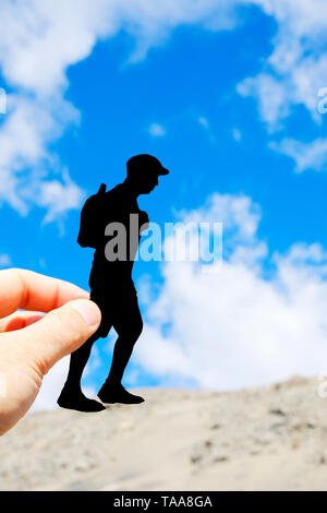 Nahaufnahme der Hand eines kaukasischen Mann hält ein Papier Ausschnitt in der Form von einem jungen Mann, trug eine Kappe und einen Rucksack, wandern durch die Dünen Stockfoto