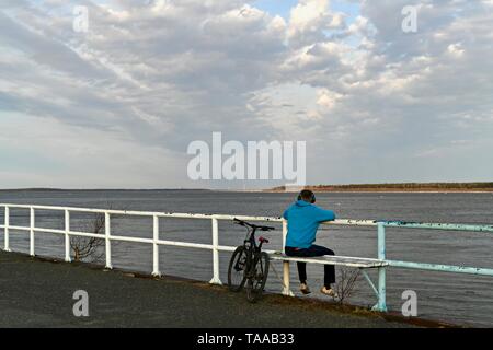 Surgut, Khanty-Mansiyskiy avtonomnyy Okrug, Rossiya - 10. Mai 2019: Damm des Ob-Fluss. Die ice Drift zu einem Ende kommt. Wolken schweben niedrig und der Himmel wird eine Reflexion auf den Fluss. Nach einem langen Winter, Bewohner des Nordens endlich ein Wechsel der Jahreszeit erlebt. Ein junger Mann mit Kopfhörern sitzt auf der Bank der Damm und in die Ferne schaut auf den Fluss, auf die Horizontlinie, wo der Fluss Himmel treffen. Sein Fahrrad in der Nähe geparkt. Stockfoto