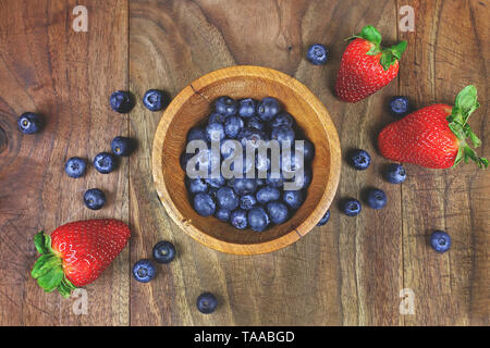 Eine kleine hölzerne Schüssel Blaubeeren wird durch frische, gesunde Erdbeeren auf einem urigen Laufbelag Hintergrund umgeben. Stockfoto